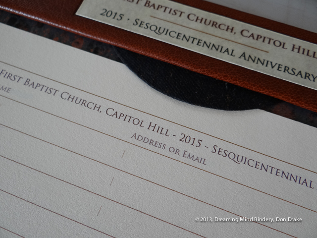 Detail of a leather and marbled paper sign-in station board showing the page hold-down tab, lacquered paper label and part of the guest sign-in page design