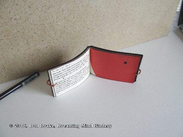 A miniature book being held open with a wire bracket to allow photographing its interior